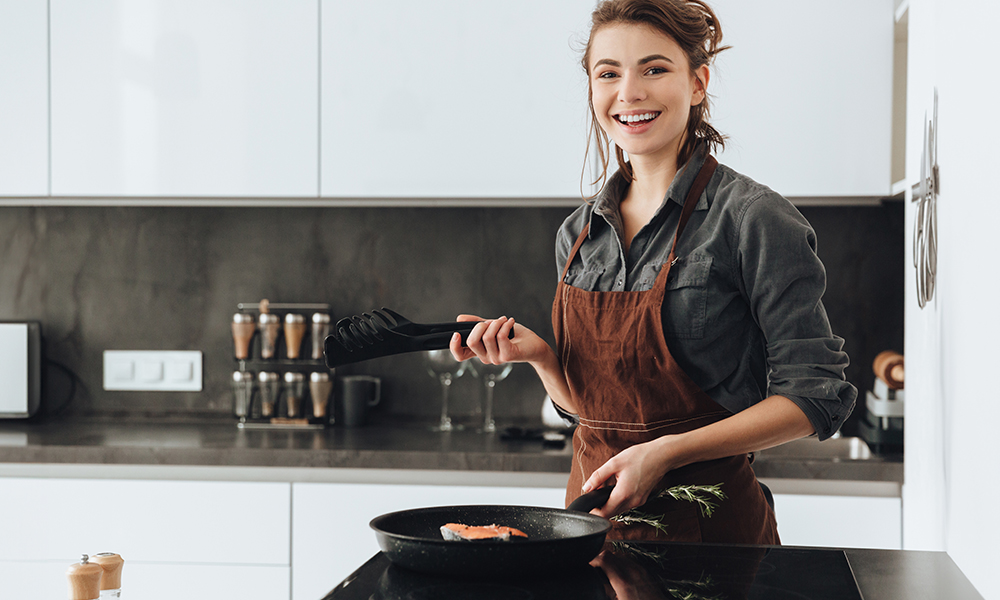 Uśmiechnięta młoda kobieta smażąca obiad na patelni w nowoczesnej kuchni