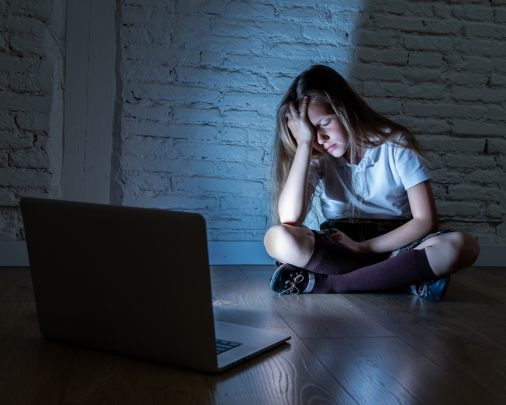 Zmartwiona dziewczynka w ciemnym pokoju, oświetlana jedynie przez ekran laptopa