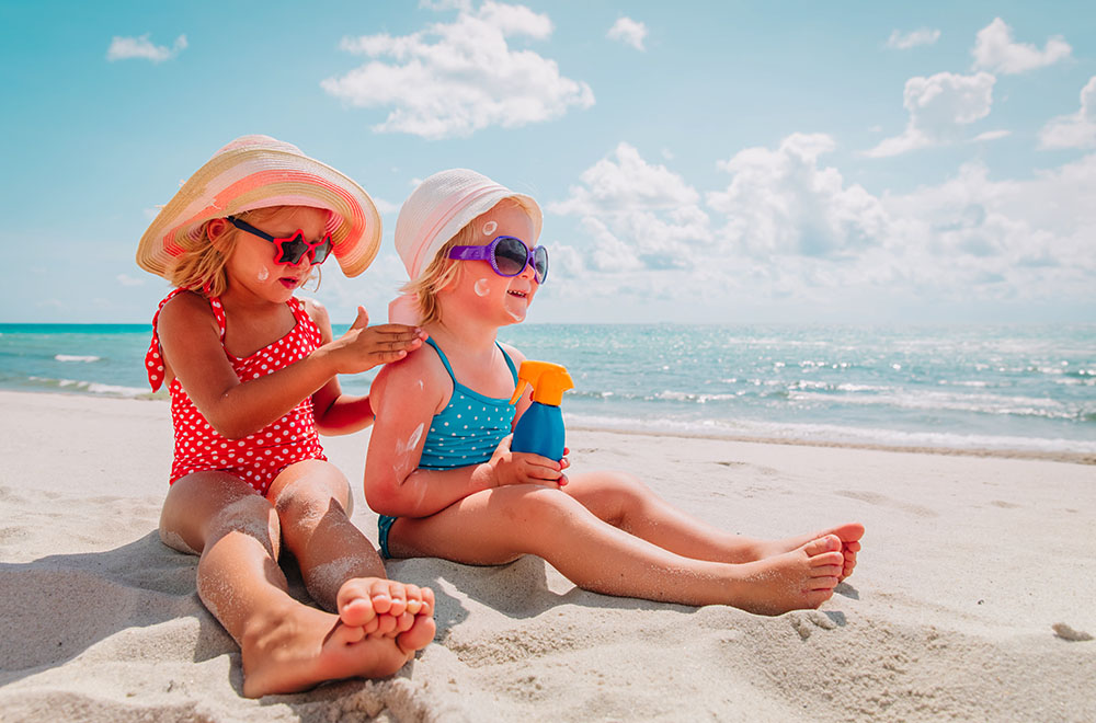 Dwie dziewczynki na plaży w strojach kąpielowych, z założonymi okularkami i kapeluszami, które smarują się kremem przeciwsłonecznym. - zdjęcie