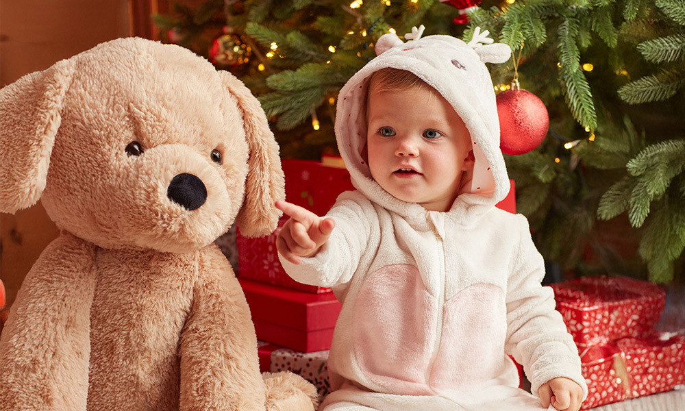 Dziecko w ubranku z reniferem siedzi obok dużego pluszowego psa, a w tle widać choinkę i prezenty. - tytułowa